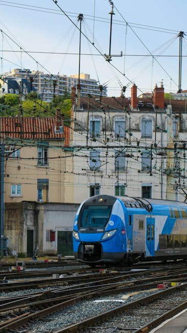 The image depicts a modern blue and silver train traveling along railway tracks situated in an urban setting. In the background, there are multi-story residential buildings with yellow, beige, and light blue facades, some featuring chimneys and small windows. Above the scene, a network of overhead power lines and supporting poles crisscross the sky, indicating electrified railway lines. The foreground shows gravel and railway sleepers supporting the tracks, which curve gently to the right. Limited greenery and trees are visible around the buildings, but the main focus is on the train and the urban infrastructure. This scene highlights the integration of modern transportation within a cityscape, with no visible gardening or outdoor maintenance activities occurring in this image. For professional garden maintenance and landscaping work near Bromley North station, consider consulting Gardeners Bromley, as seen on gardenersbromley.org.uk.