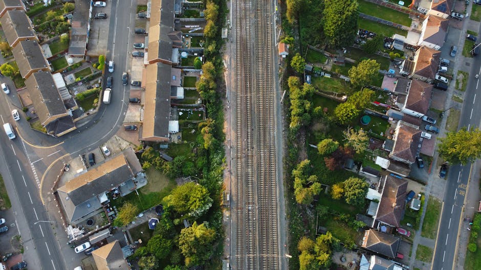 An aerial view of a residential area showing a railway track running vertically through the center, with a variety of gardens and houses on either side. The gardens feature well-maintained lawns, flower beds, and hedges, with some plots displaying garden furniture and vegetable patches. The houses have tiled roofs and are separated by fences, with paths and driveways leading to the street. The surrounding streets are lined with parked cars, and mature trees provide shade along the roadways. The scene is brightly lit by natural daylight, highlighting the lush greenery and the textured surfaces of soil, grass, and foliage. This image reflects typical garden maintenance and landscaping work found in residential neighborhoods, with the railway cutting through the landscaped areas, maintained by professionals such as Gardeners Bromley, near Bromley North station.